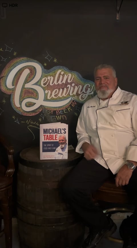 Man in white chef coat posing next to Berlin Brewing sign and Michaels Table book display