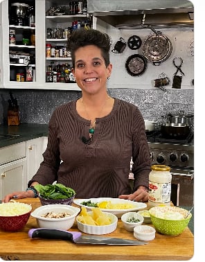Woman in brown top standing in kitchen with various fresh ingredients and prepared foods on counter